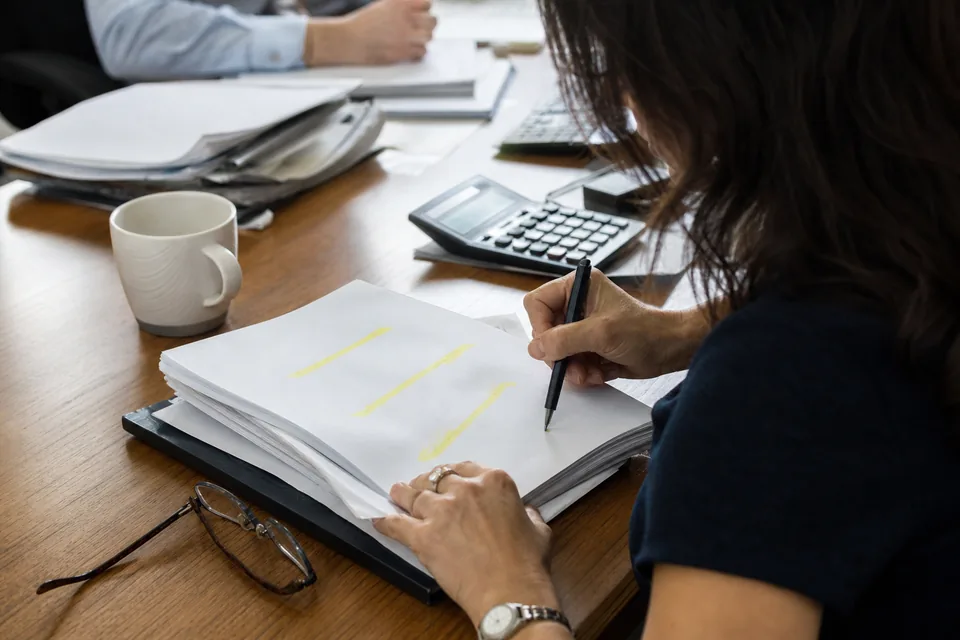 Hand annotating highlighted pages of a price schedule beside a calculator on a desk